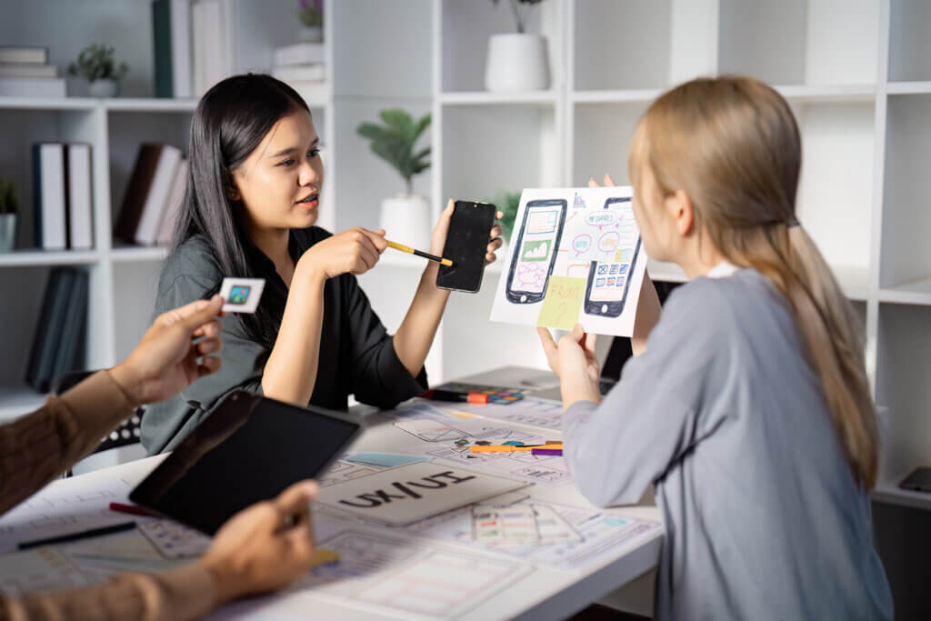 Asian UX designer discusses mobile UI design with mobile app prototype in hand, as dev white woman listens.