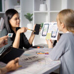 Asian UX designer discusses mobile UI design with mobile app prototype in hand, as dev white woman listens.