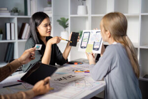 Asian UX designer discusses mobile UI design with mobile app prototype in hand, as dev white woman listens.